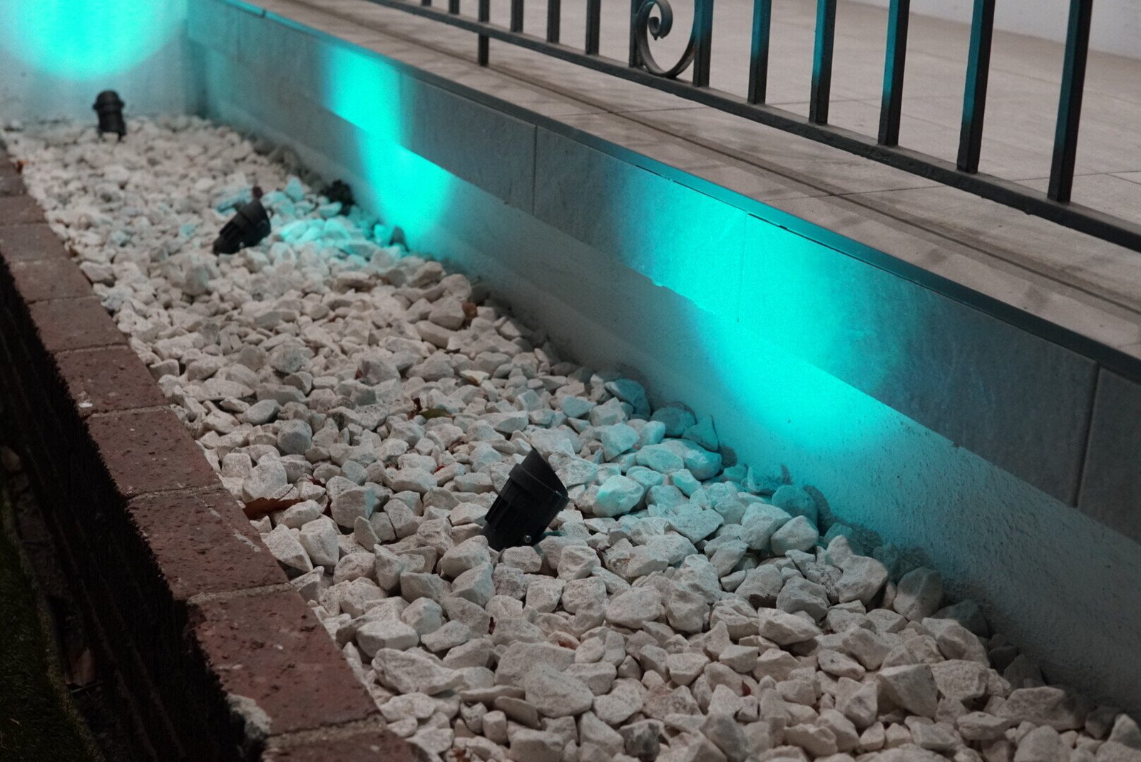 Outdoor landscape lighting installed in a bed of white decorative rocks, casting a teal glow against a low wall with a black iron railing.