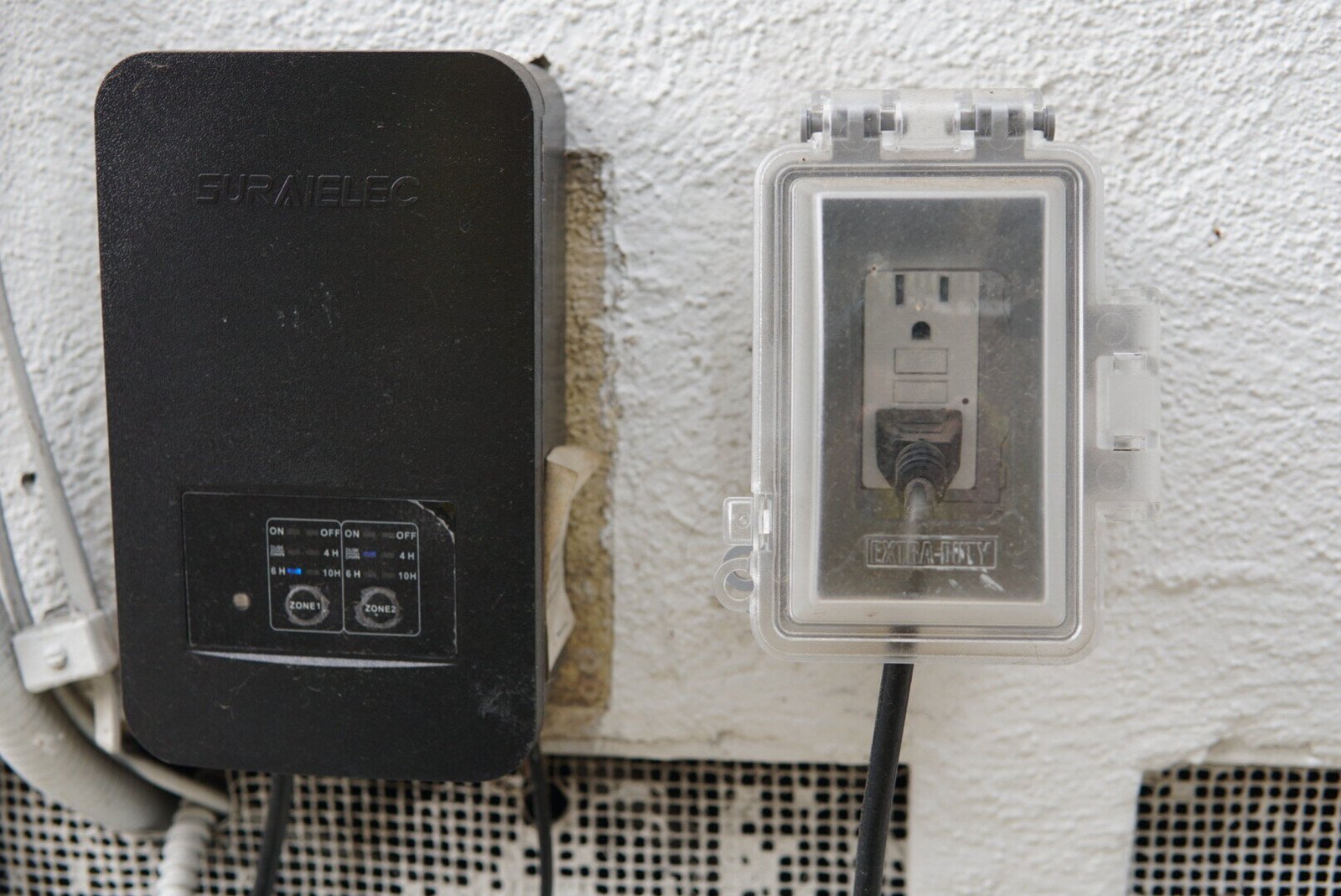Outdoor electrical outlet with a protective cover installed next to a black electrical control box mounted on a white wall