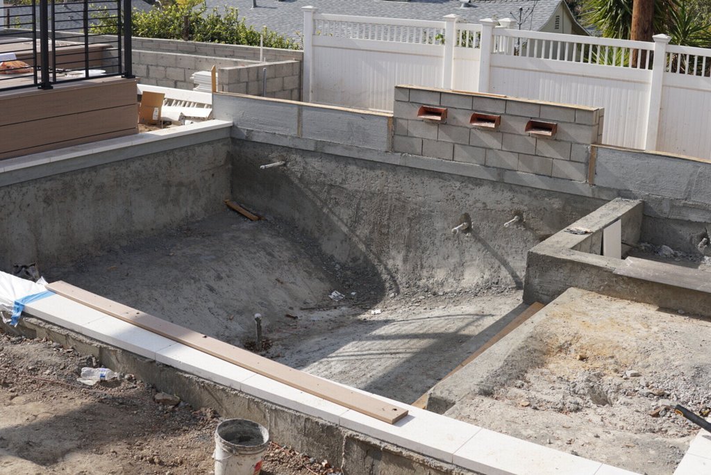Under-construction swimming pool with exposed concrete structure, plumbing pipes, and surrounding retaining walls.